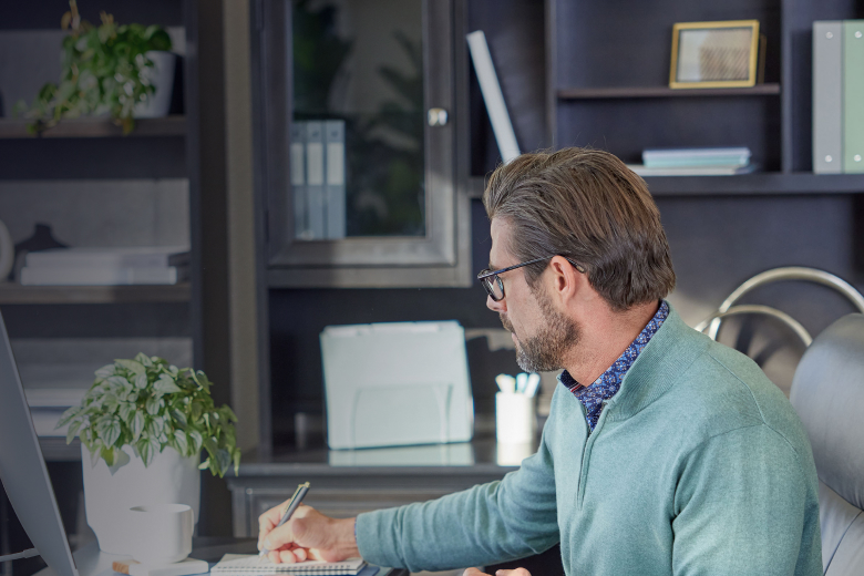 A young man in a black T-shirt sits at a foldable desk with a laptop and a coffee cup, looking at the camera. To the left, large white text reads “Need better office comfort? Let’s solve that.” A potted plant is on the right, and the background shows a bl